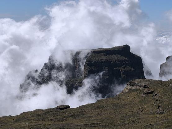 Clouds are sucked up vertically in the air streams and engulfs a large blocky peak.  The right side of the peal is visible, but the left-side is obscured in places by the cloud. There are some cloud above the peak.
