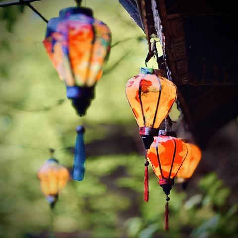 Warm, colorful lamps with leaflike patterns hang in a "J" formation from awnings beneath several trees on a cloudy day.