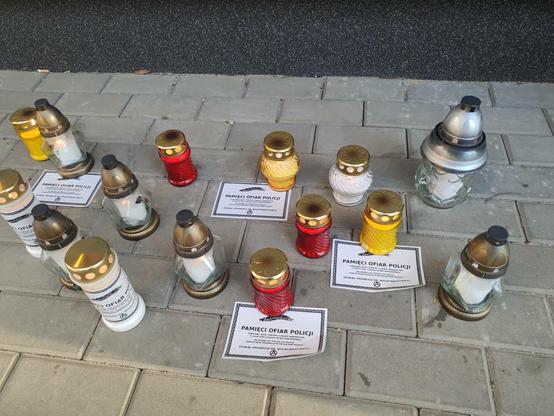 Zdjęcie przedstawia znicze na tle kostki brukowej. Pod niektórymi jest klepsydra z napisem "pamięci ofiar policji"