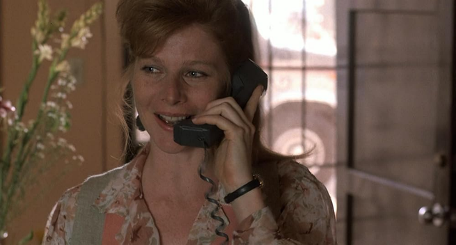 Actress Lisa Moncure is shown in a still from the movie, "Lisa (1990)."  She is in an interior location, there is a window behind her and flowers in a vase next to her.  She appears to be standing, but shown from the shoulders up.  She has a black corded phone receiver help up to her ear and looks cheerful.  She is wearing a colorful patterned blouse and her hair is up.