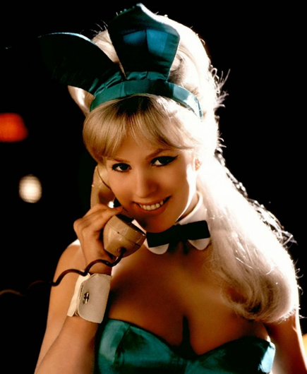 Model June Havoc is wearing a green Playboy bunny outfit complete with green ears and bow tie.  She is shown from the bust up on a dark background that a red and white point light on it.  June is looking straight at the camera and smiling slightly.  A strong high light comes from one side on her long, striaght,  blond hair, which falls down over her shoulder.