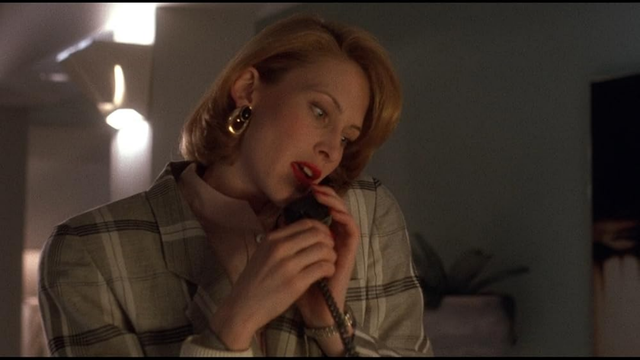 Actress Frankie Lou Thorn in a still image from the movie, "Lisa (1990)."  She is in an interior location,  sitting on a plaid couch that, oddly, almost matches her jacket.  There is a lamp in the background that is apparently lighting the room.  She is shown from the waist up, wearing a brown plaid jacket.  She has a phone receiver up to her ear that she is holding with both hands.  A large gold earring is hanging from her other ear.  Her shoulder length hair is styled as a bob.