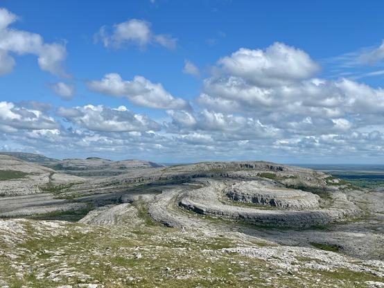 A barren, treeless landscape reveals a hill composed of swirling layers of limestone rock. Scatter grasses and rocks in the foreground; puffy white clouds in a light blue sky above.
