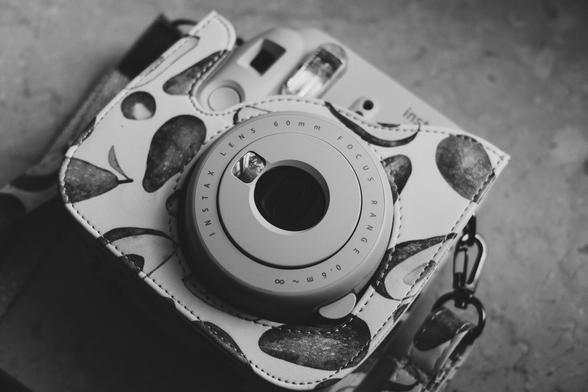 Black and white close-up of a Fujifilm Instax instant camera, partially enclosed in a patterned leather case with an avocado design. The camera’s 60mm lens and flash are visible, emphasizing texture and contrast in the image.