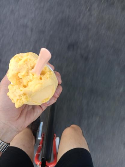 A view looking down. There is a hand holding a cup with yellow ice cream and a spoon. Below that you can see a pair of legs and the wheel of a unicycle.