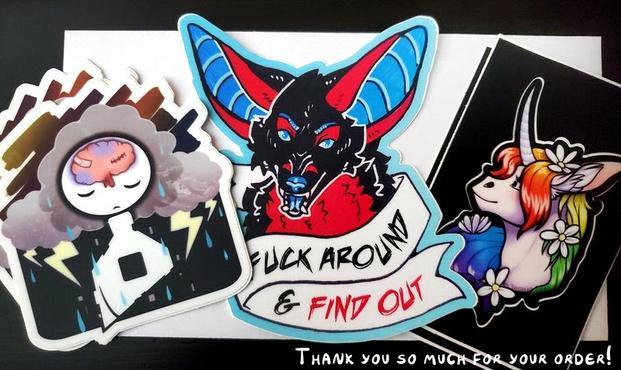 A photo of stickers on top of a white envelope, sitting on top of a black surface. Far left shows a mental health sticker of a simple shaped human figure with a black hole in their chest, a bruised brain, and rain clouds with lightning around their head. Above that where there should be clear sky, is muted colours and foggy. Middle sticker is a black, red, and blue canine creature snarling with a ribbon that reads "Fuck around & find out". On the right is a unicorn head with rainbow mane flowing around it, flowers in their hair and they are smiling a little while facing the left.