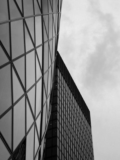 Black and white photo looking across the curtain wall façades of a pair of office buildings. The structure on the left features a curving surface composed of triangular and diamond shaped panes of glass. The tower on the right exhibits a flat wall made up of rectangular windows set into a metal framework.