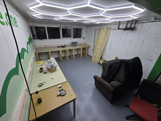 Ein Bild von dem neuen 4. Raum im vspace.one. Der Boden ist Grau und von der Decke beleuchtet ein sechseckiges Lichtmuster den Raum. Die Wände sind Weiß und auf der linken Wand ist zusätzlich eine grüne Rakete mit dessen Abgasen als Wolkenmuster aufgemalt. Außerdem lässt sich ein ebenfalls grüner vspace.one Schriftzug erahnen. Hinten befindet sich unter einem Kabelkanal mit Steckdosen und Netzwerkdosen eine Arbeitsplatte aus Holz mit einer Eurobox und Filamentrollen drauf. Darüber ist eine Reihe an Fenstern. Rechts an der Wand, bei der die Backsteine sichtbar sind, sind Holzbalken angelehnt und es sind Leitungen zu sehen. Links vorne ist ein Tisch mit einer beigen Platte, auf dem sich verschiedene Werkzeuge befinden. Rechts liegen zwei braune Sessel aufeinander. Davor steht ein Drehstuhl.