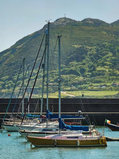 Several small sailing boats with masts are moored in calm, blue water alongside a dark grey harbour wall. In the background, a large, green, undulating hill rises up to meet a clear, pale blue sky. A cross is visible at the top of the hill.