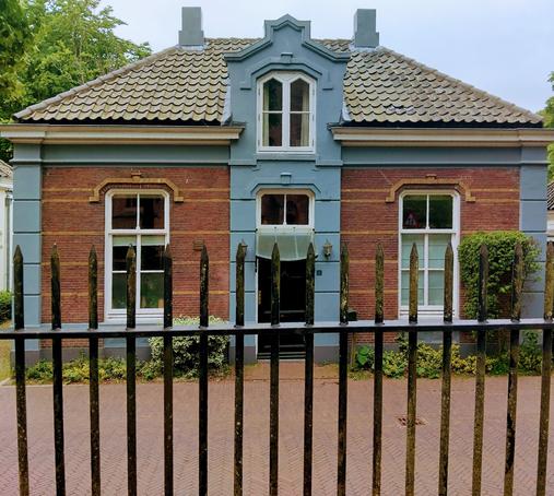 Facade of brick house behind a fence with an ornate stone entry with a door and a window above. ¦ Façade van een bakstenen huis achter hekwerk met een geornamenteerde stenen ingangspartij met een deur en een raam erboven.