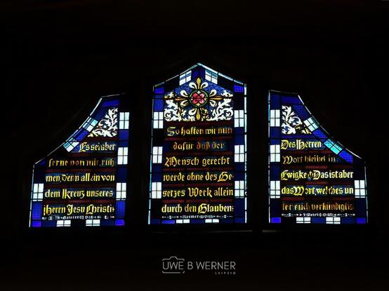 Kirchenfenster in der Schlosskirche Wittenberg mit biblischen Inschriften auf Deutsch, gestaltet in kräftigen Blau-, Weiß- und Goldtönen mit kunstvollen Verzierungen.
