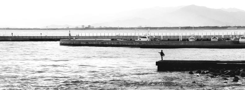 The image shows a black and white scene of a waterfront with a calm body of water, possibly a sea or lake. There is a person standing on a pier or dock extending into the water, possibly fishing. The background includes a barrier, some boats docked along the edge, and distant mountains. The atmosphere is tranquil and serene.