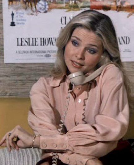 In a scene from the "Rockford Files" Priscilla Barns is on the phone, perhaps in an office setting.  Behind her is a large movie poster, which is only partially visible.  She is wearing a mauve blouse and is holding the phone between her shoulder and ear, with her hands crossed in front of her.  Her long blond hair is hanging down her back.