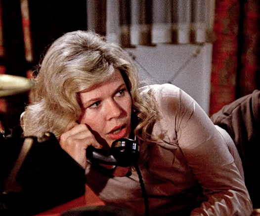 Loretta Swit in the TV show M*A*S*H.  She is in an interior setting, probably her tent.  She is leaning forward from a chair onto a table where there is a telephone, she has the receiver up to her ear.  She is wearing a khaki military shirt and is looking away from the camera.  Her mouth is open, apparently talking on the phone.  Her silvery blond hair is in a flip style around her shoulders.