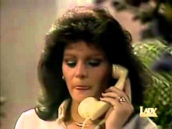 I have not identified who this actress is, but this a still from "Heart of Stone."  She is shown from the shoulders up in an interior setting, with greenery behind her.  She is holding a yellow phone receiver and her eyes are almost closed as she seems to be listening intently to the phone conversation.  She is wearing a white blouse and has a very large wedding ring on her hand.  Her hair is shoulder length and layered and volumized.