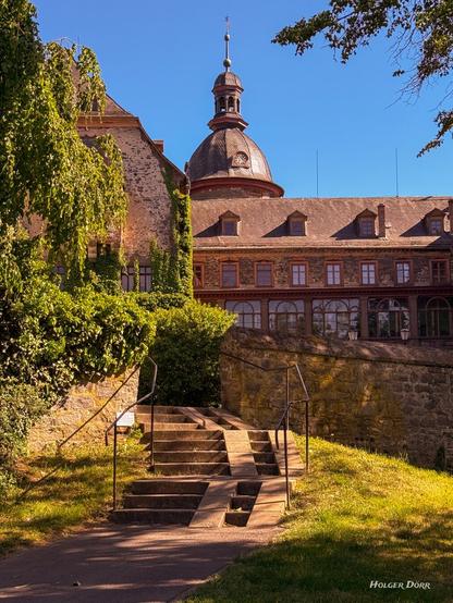 Ein steinerner Treppenweg führt durch grünes Gebüsch und Schattenlicht hinauf zum Schloss Laubach. Im Hintergrund ragt der barocke Kuppelbau des Schlosses über eine lange Fassade mit vielen Fenstern. Das Bild ist von unten aufgenommen und vermittelt eine ruhige, frühsommerliche Stimmung.