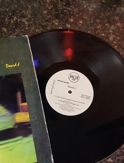 The 12" single of "I'll Be Your Chauffeur" by David J is three-fourths of the way out of its sleeve. Red and blue light is reflecting off the black vinyl. The visible portion of the cover shows "David J" written in yellow cursive on a grey/green background.