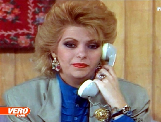 "The Revenge" was a Venezuelan telenovela from 1989.   Carmen Julia Álvarez is shown in a still image from the show.  She is in an interior location with a paneled wall and wall hanging behind her.  She is holding a two toned, blue and white midcentury modern phone receiver to her ear and looks straight at the camera.  Her head and shoulders are visible, she is wearing a light teal jacket with a dark blue blouse underneath.  She is wearing far too much makeup.  Her red hair is very styled, layered and sculptured.