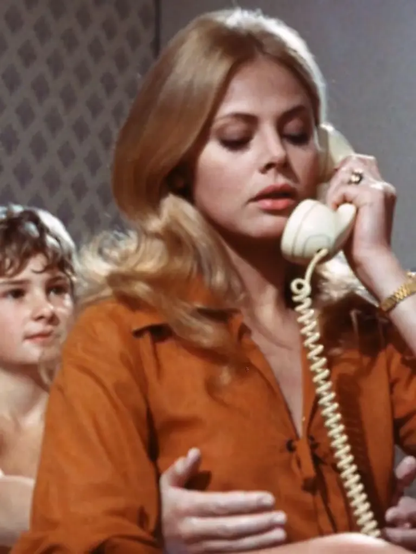 In a still from the movie, "What the Peeper Saw," Britt Eklund is sitting on a bed, with Mark Lester sitting in the background behind her.  She has a white corded phone receiver held to her ear and is looking down, looking like she is concentrating.  From behind, Mark has his hands around Britt, almost cupping her breasts.  She is wearing an orange blouse and her blond, loosely curled hair hangs down below her shoulders.