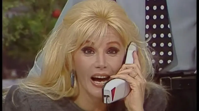 Venezuelan talk show host Susana Gimenez is talking on the phone.  A man is standing behind her, only his torso and tie are visible.  Her mouth is wide open and she looks either surprised or is yelling at the person on the phone.  She is wearing a gray suit and only her head and shoulders are visible.  She has straight platinum blond hair which falls down to her shoulders.