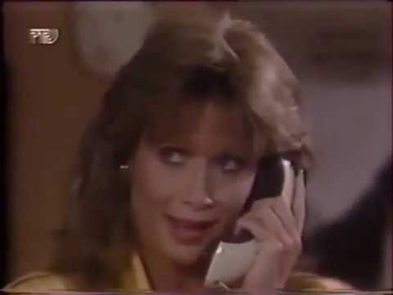 Julie Condra is in an interior setting, with and indistinct background.  Only her face and the top of her shoulders are visible.  She is holding a white and black phone receiver that has a curved, modern shape.  She seems to be wearing a yellow shirt.  Her eyes are averted to one side.  Her hair is shoulder length in a shaggy, layered style.