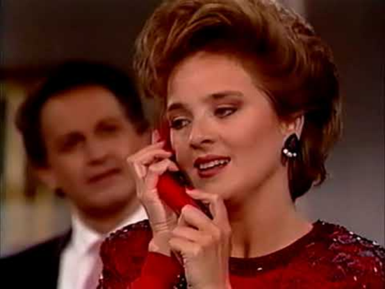 Barbara English is standing in an interior setting, although only her shoulders and face are visible.  A man in a black jacket and white shirt is standing behind her.  She is wearing a red dress and is holding a phone receiver with both hands to her ear.  Her hair is in an elegant layered updo.