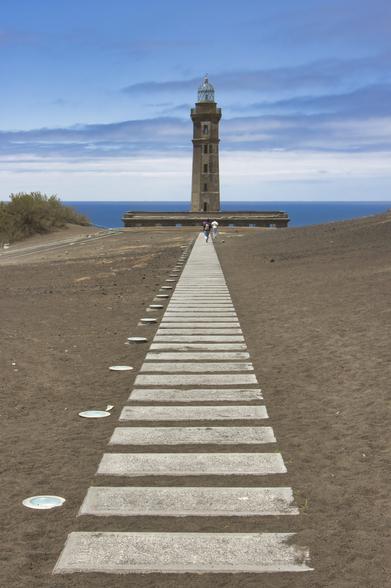 Blocchi di cemento incastonati nel terreno che conducono ad un grande faro all'orizzonte.