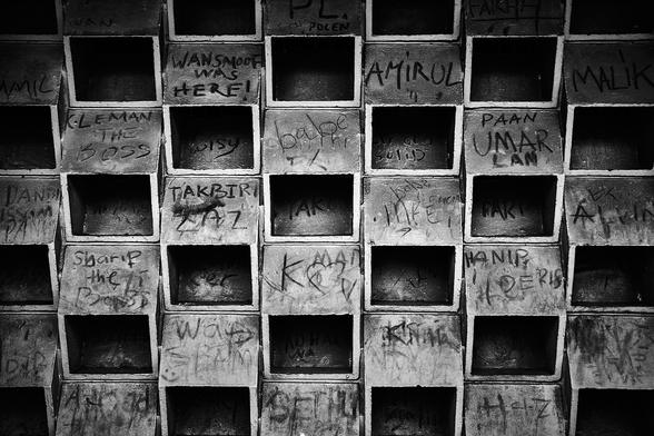 A black and white image of a building's wall with a grid of small, square openings. Many of the spaces between the openings are covered with various scribbles and signatures made by different people.