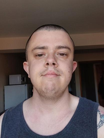 A white transmasc with a buzzed head, and stubble. He's in his flat and is wearing a grey vest