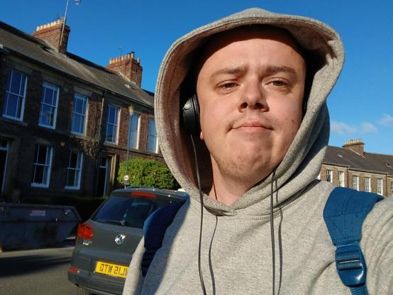 A white transmasc wearing a grey hoodie, a blue rucksack, and black headphones. He's stood in front of a row of edwardian houses.