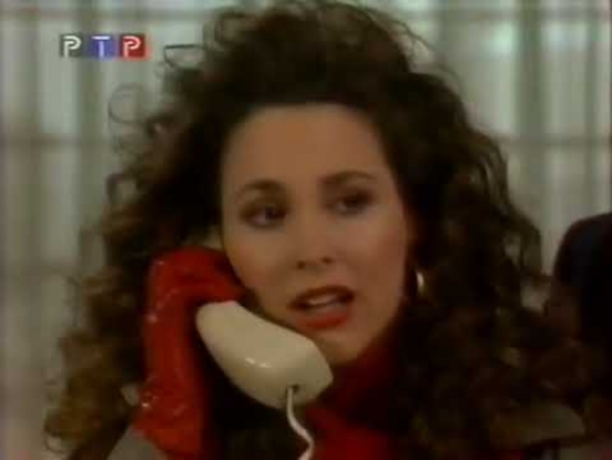 Karen is in an interior location with a window covered with translucent drapes behind her.  Her head and shoulders are visible, with her hand in a red glove, she is holding a white princess phone receiver.  She is facing the camera, wearing a black jacket with a red shirt underneath.  Her long dark hair is extremely curly and comes down to her shoulders.