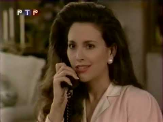 Karen is in a brightly lit interior, a white chair or couch in the background.  She is sitting, head and shoulders visible.  She is wearing a light pink sweater or jacket.  In one hand she has a black phone receiver up to her ear, facing the camera.  Her dark hair is straight and falls below her shoulders.