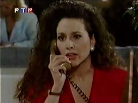 Karen is sitting in an interior setting, possibly a restaurant, there is a person sitting in the background behind her.  Her head and shoulders are visible, she is wearing a red V neck dress.  She is holding a light brown phone receiver up to her ear and has a look of concentration on her face.  Her dark hair looks lightly permed and falls down over her shoulder.