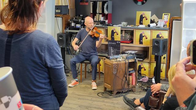 Der Künstler mit Violine und ein wenig elektronischem Equipment wie Mischpult und Looper. Hinter dem Künstler seine neue Platte, goldenes Cover, mehrfach aufgestellt.