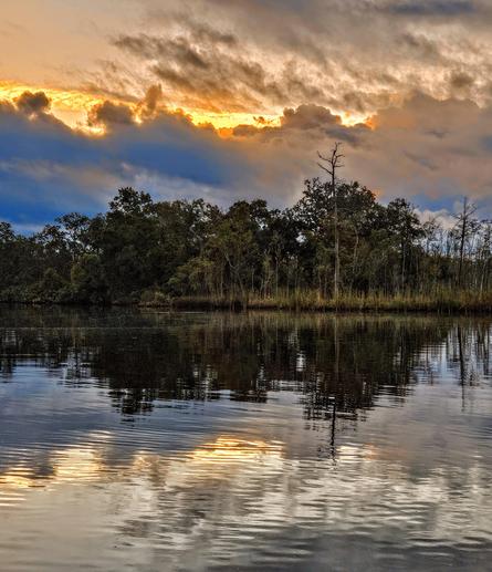 Firey, explosive colors rise from beyond blue storm clouds rolling over a lush wetland peninsula and waterway,  casting reflections upon the water below.