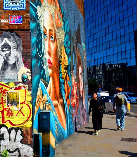 Bright street art featuring a woman's face covers a brick wall; two people walk past on a sunny, urban sidewalk near a glass building.

Ein helles Strassenkunstwerk mit einem Frauengesicht bedeckt eine Backsteinmauer; zwei Menschen gehen auf einem sonnigen, staedtischen Buergersteig in der Naehe eines Glasgebaeudes vorbei.