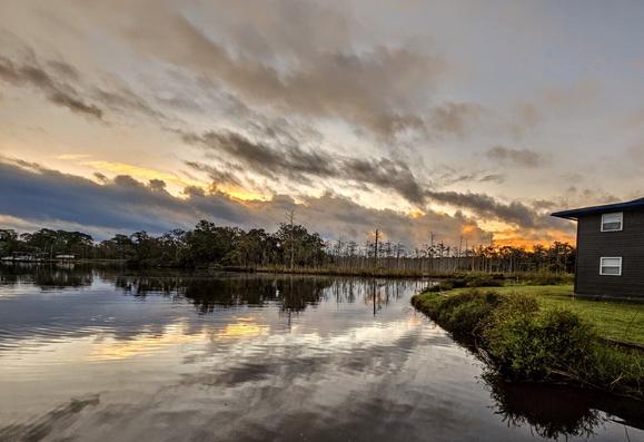Firey, explosive colors rise from beyond blue storm clouds rolling over a lush wetland peninsula and waterway,  casting reflections upon the water below.