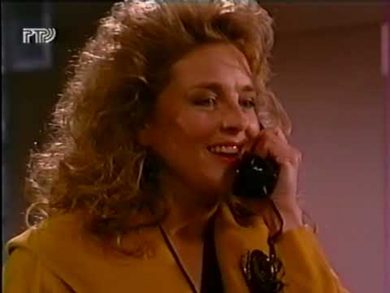 Robin Mattson is in an interior location with an indistinct light gray background.  She is shown from the shoulders up, wearing a yellow/orange jacket, a similar color blouse and a black tie.  She has a black phone receiver to her ear with a pleasant look on her face.  Her curly, blond, possibly permed hair, falls just down to her shoulder.