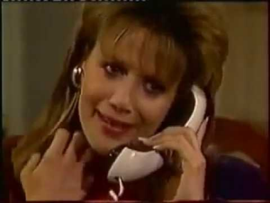 Julie Condra is in an interior location in a close up shot, showing just her shoulders and face.  She is sitting on a red chair.  She has a white and black phone with a modern curved design up to her ear.  Her other hand is up on her shoulder, fingers extended.  She has a slight smile on her face and her hair is a bit of a female mullet, curled and volumized on top and straight down the sides to her shoulders.