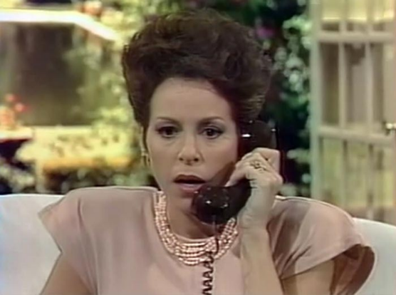 Louise Sorel is in what looks like an outdoor location, perhaps a patio.  There is indistinct lattice works and greenery in the background.  She seems to be sitting, but only her head and shoulders are visible.  She is wearing a pink/mauve dress with a ringed collar and enormously wide shoulder that are open underneath.  She has a black phone receiver in her hand, up to her ear.  She has a concerned look on her face and her mouth is slightly open.  Her tightly permed hair is shaped into a dome over the top of her head.