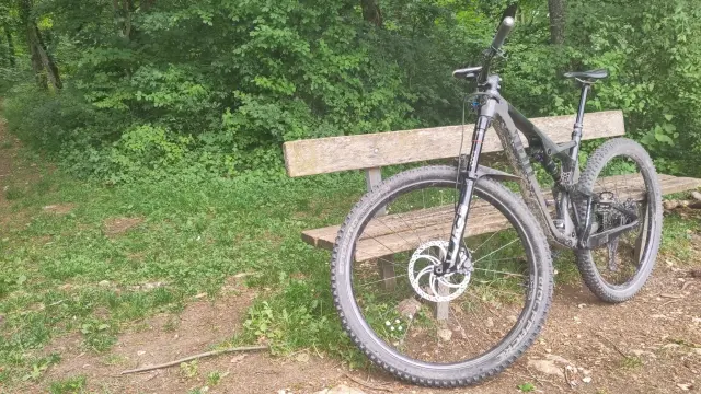 Ein Mountainbike lehnt an einer Sitzbank. Im Hintergrund sieht man Wald und einen schmalen Weg (Trail)