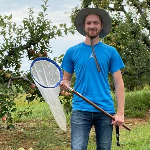 The photo shows a man (Robert Ostrom) in a blue shirt, jeans and a hat. He holds a insect net across his body. He is smiling and looks ready to collect some insects.