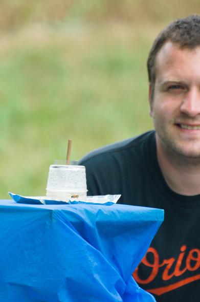 An inverted small mason jar is placed on a blue table cloth on a tripod. The mason jar serves as a sugar syrup feeder for bees. A man (Brad Ohlinger) with dark hair and a black shirt sits behind the tripod to observe honey foraging. He is slightly out of focus, but candidly looks at the camera.