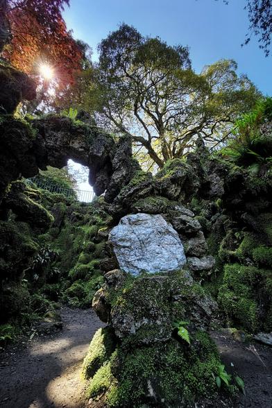 A low-angle shot shows a sunlit, moss-covered rock archway and surrounding rocks, with a large, pale, textured stone in the foreground and a tall tree reaching towards a bright blue sky in the background. Sunlight flares through a red-leafed tree at the top-left.