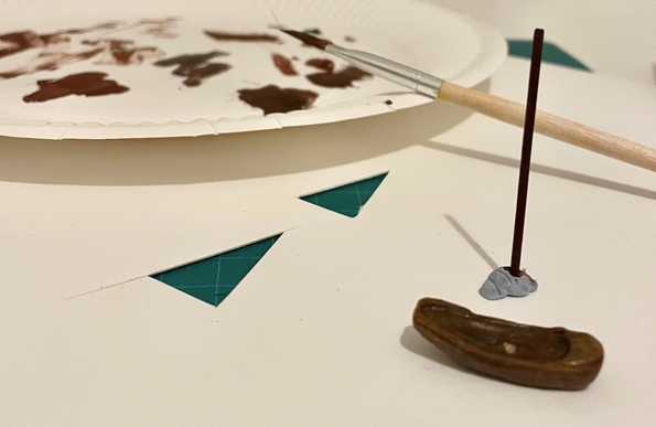 Image description by Tiny Crew Big Raffle: A hull of of Tiny Crew boat, on a white sheet of card with two triangle flag shapes cut out. A painted mast is behind the hull, with a palette and a paintbrush covered in brown paint behind that.