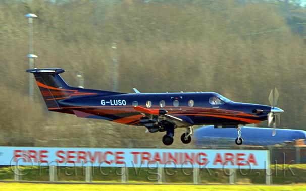 Side view of a black, single engined BizProp flying from left to right at a very low altitude, with undercarriage lowered and flaps extended from the rear of the wings, suggesting it is about to land.
The plane is black, with red stripes swooping from under the nose, over the wing, and back under the rear fuselage, and more on the tail.
The white registration "G-LUSO" is on the upper rear fuselage.
In the background, a white banner with red text "Fire Service Training Area" stretches across most of the frame from the left.
The blue fuselage of a fire training rig is visible in the background under the nose of this plane, with trees filling the rest of the background.
