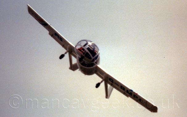 Low, almost head-on view of a white, single engined light aircraft with a bulbous, heavily glazed cockpit canopy in front of a fuselage that is basically a shrouded propellor engine in the middle of a long, straight wing.
There are long tail-booms extending part way through each of the wings, with the fixed undercarriage attached to the front of the booms.
The plane is heavily tilted to the side, obviously performing a spirited flying display routine.
Grey sky fills the rest of the frame.
