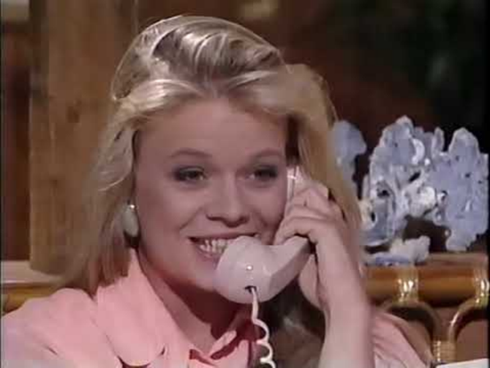 Marcy is an interior location, with a bookcase with a sculpture behind her.  Her head and shoulders are visible, wearing a pink shirt.  She is holding an off white phone receiver to her ear, she has an eager expression on her face.  Her blond hair is straight and falls to her shoulders.