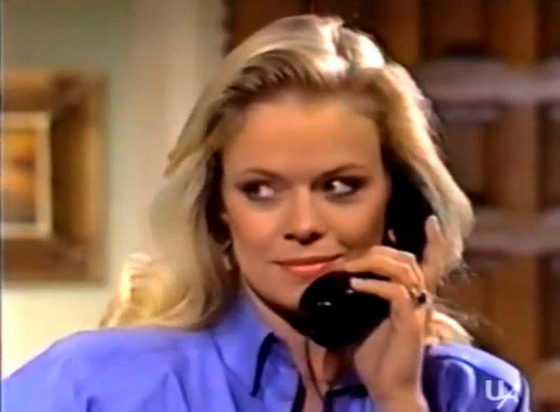 Marcy is in an interior location with some kind of wood lattice directly behind her and a wall with a framed picture on the other side.  Her head and shoulders are visible, she is wearing a bright blue blouse.  She is holding a black phone receiver to her ear and giving a definite side eye, with a bit of a smirk on her face.  Her straight blond hair is down behind her shoulders.