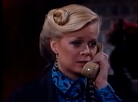 Marcy is in a dark interior setting, with an indistinct background.  She is standing, but only her shoulders and face are visible.  She is wearing a black jacket with a blue polka dot blouse underneath.  She is holding a beige phone receiver to her ear.  Her hair is tightly styled to her head, with a swirl on one side.